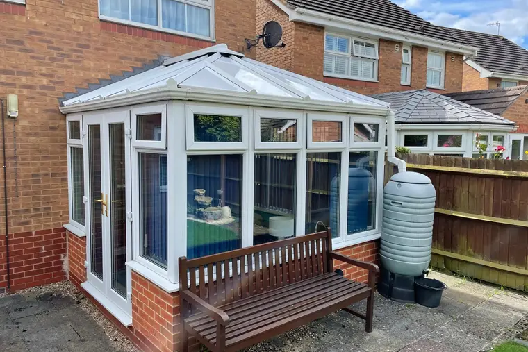 White Victorian conservatory with a new insulated panel roof beside a garden bench and water butt
